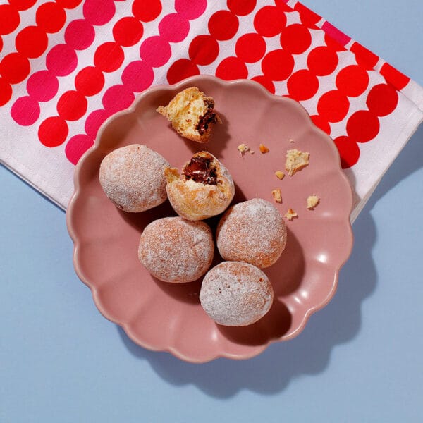A plate, viewed from above, containing six of our delicious Mini Chocolate Hazelnut Beignets. Two of the beignets have been opened and the filling can be seen inside.