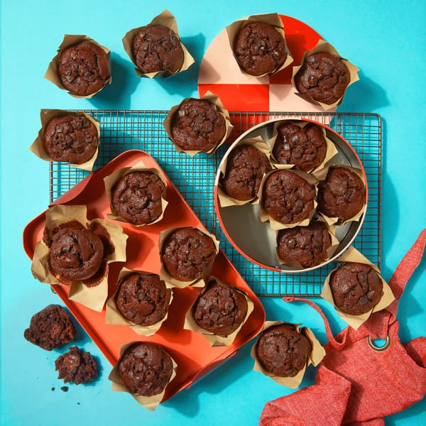 A full case of Chocolate Muffins by Cake Club on a tray and cooling rack with a light blue background.