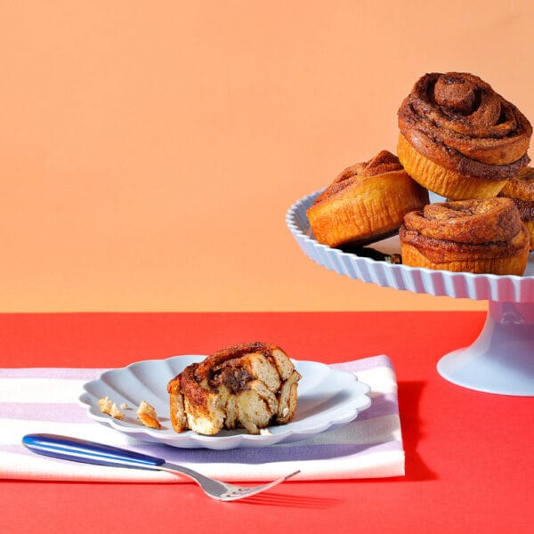 A sliced cinnamon bun on a plate with a stack of cinnamon buns on a cake stand on a red and peach background.