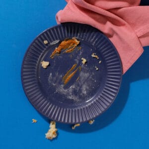 An overhead view of the crumbs from speculoos mini beignets on a ridged dark blue plate sitting on the edge of a pink ruffled napkin on a blue background.