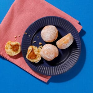 An overhead view of four speculoos mini beignets on a dark blue ridged plate with one beignet ripped open on a pink napkin on a light blue background.