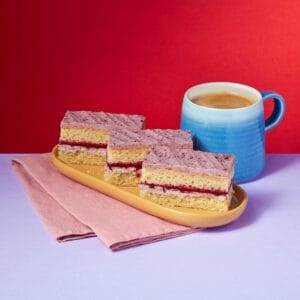 Blackberry and Mascarpone Traycake slices stacked beside a blue coffee mug on a red background.