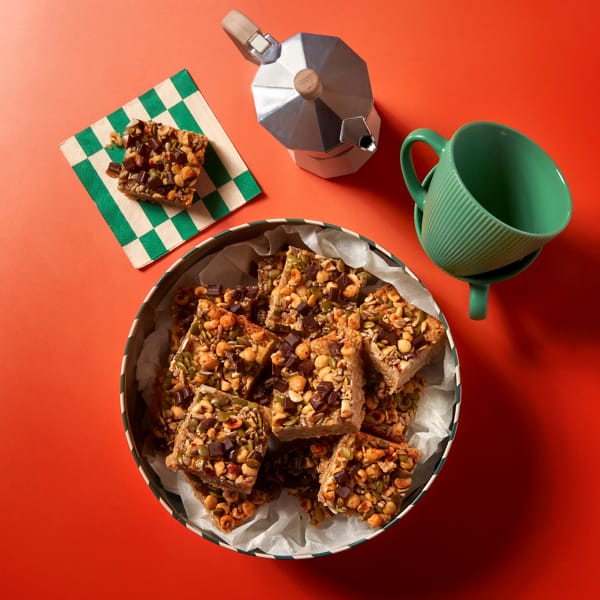 A full tray of Roasted Hazelnut Flapjacks by Cake Club on an orange background next to a green coffee cup.