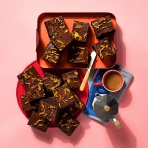 A whole tray of vegan Chocolate Orange Joy on orange and red plates, with a mug and moka pot on a blue and grey striped cloth.