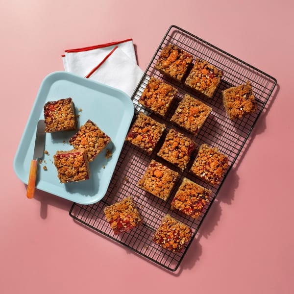 A whole tray of vegan berry crumble on a cooling rack and a light blue plate on a dusty pink background.
