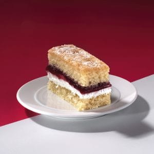 Close-up of Victoria Sponge Traycake slice with jam and cream layers on white surface.