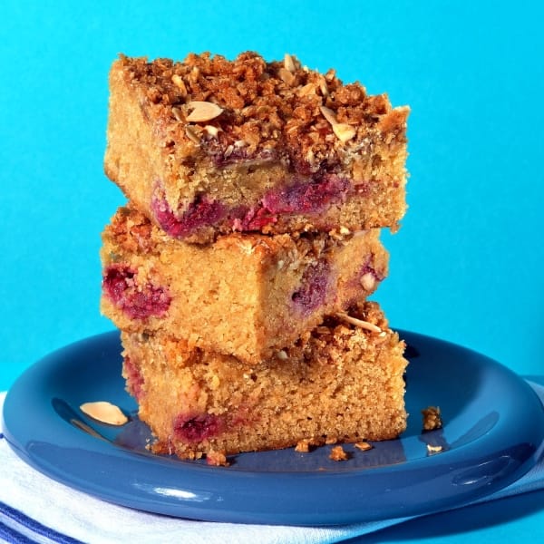 Stack of Raspberry, Ginger and Almond Traycake slices on blue plate showing layers of sponge, raspberry fruit and almond crumble topping.