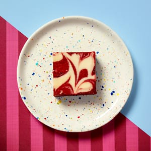 An overhead view of Cake Club's delicious Red Velvet Brownie on a white plate with multicoloured spots on a claret and light blue diagonally contrasting background.