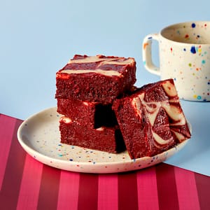 A stack of Red Velvet Brownies on a spotted plate next to a coffee cup on a claret and light blue background.